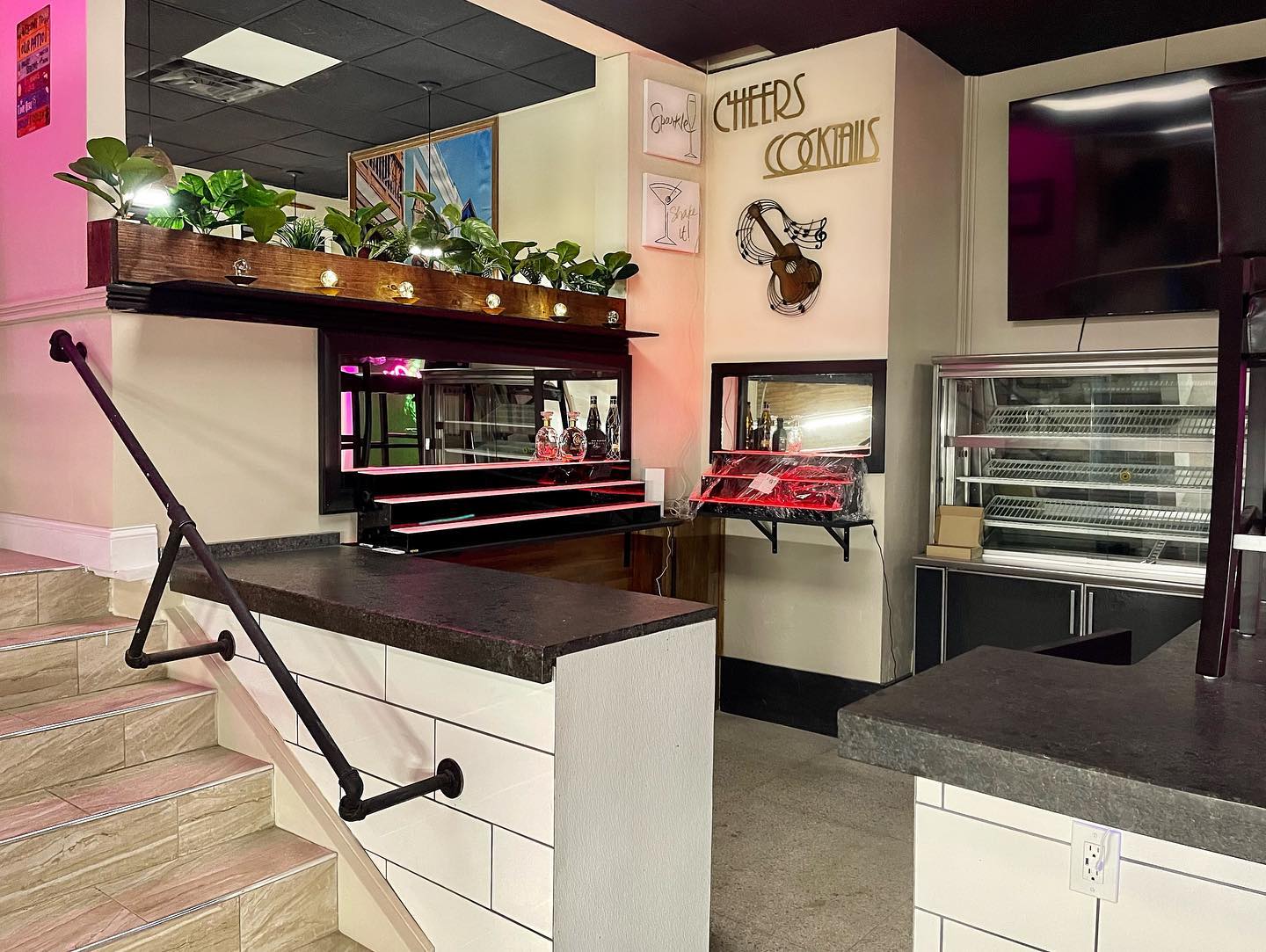 The Caribe bar with Cheers & Cocktails gold lettering, plants, and pink neon glow