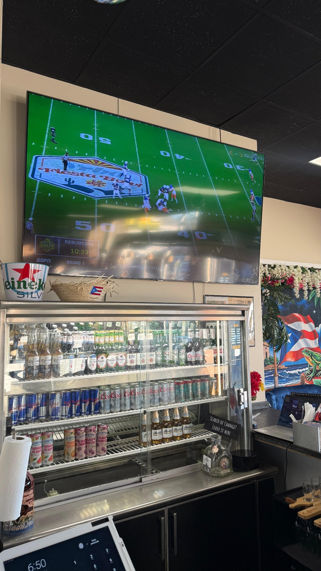 Large flat screen TV at the Caribe bar showing live college football above the cooler stocked with imported beers